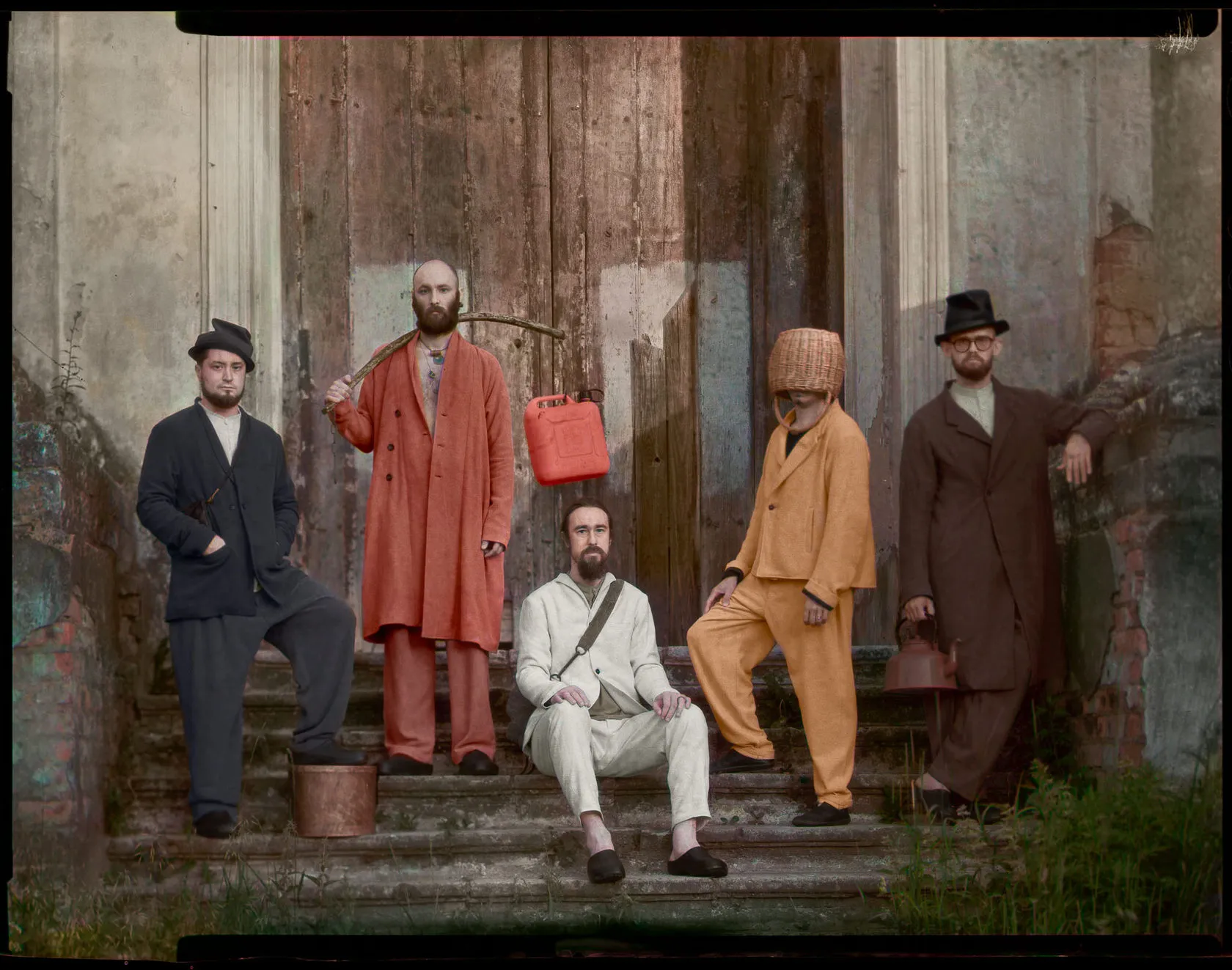 Five men pose on the worn steps of an old building, one with a wicker basket over his head.