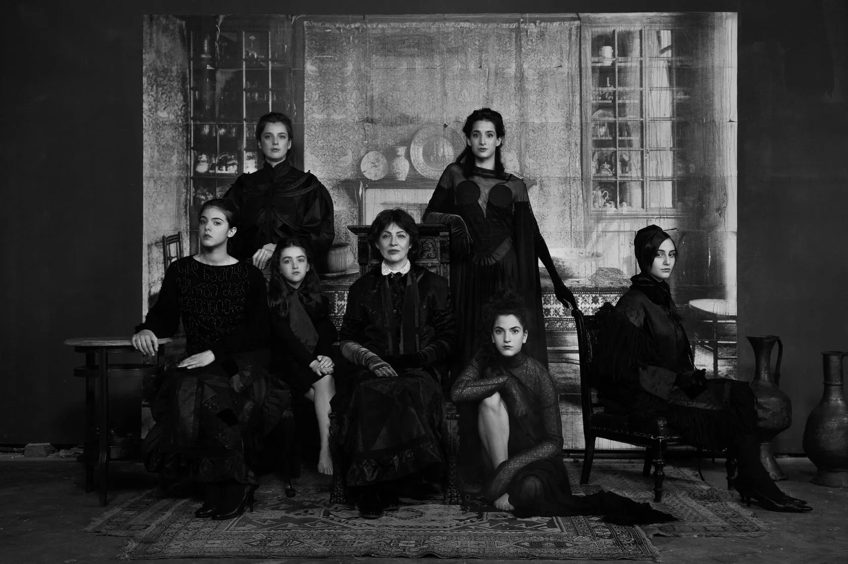 Seven women and girls in black pose before a painted room, in black and white.