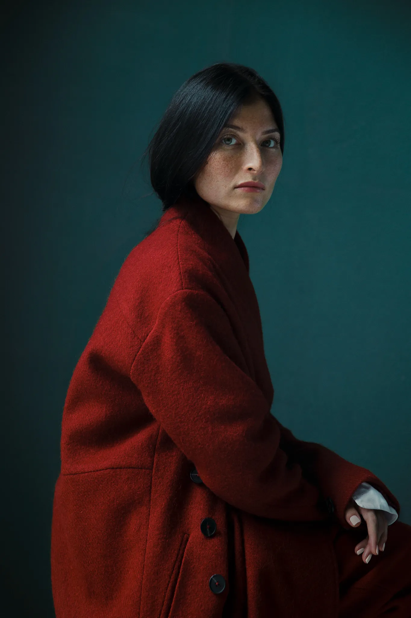Kate Brook in a red coat sits against a dark teal wall.