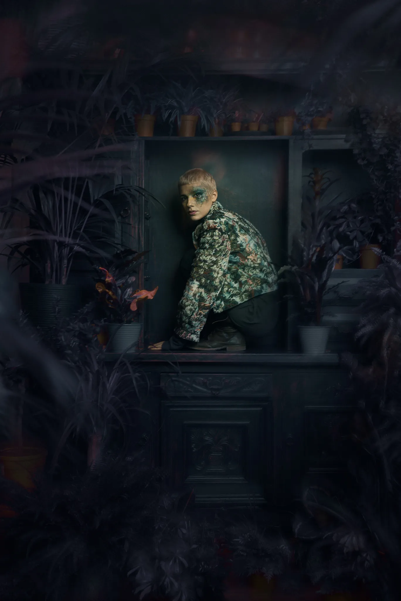 A model in a floral jacket crouches in a dark niche among plants.