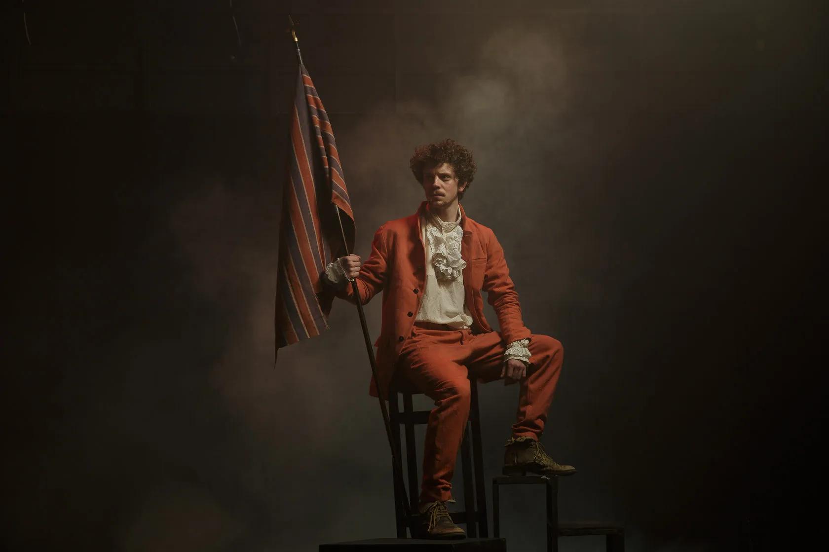 A man in an orange suit sits on a chair in drifting smoke, holding a striped flag.