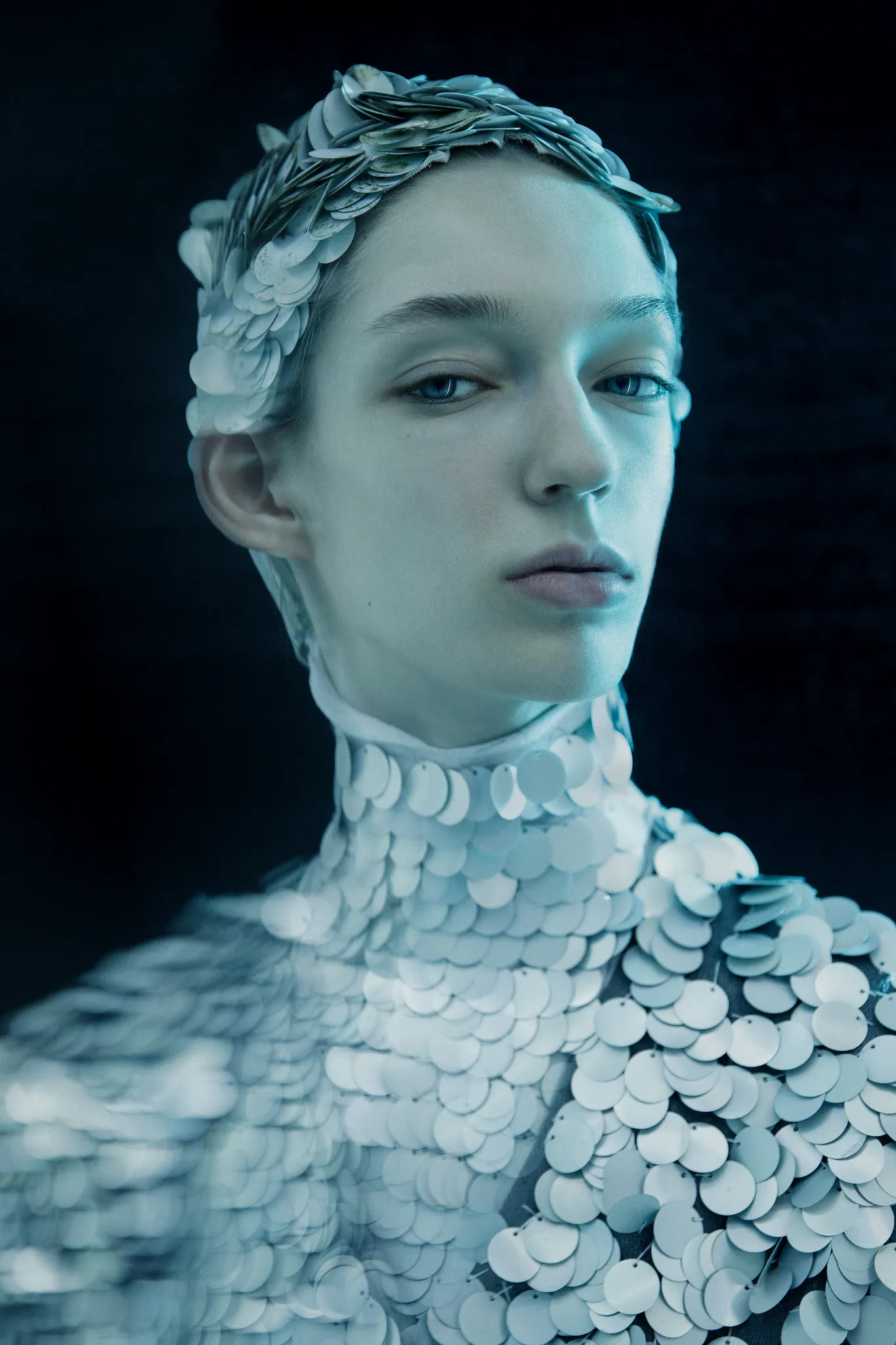 A model wears a headpiece and collar of silver sequin scales.