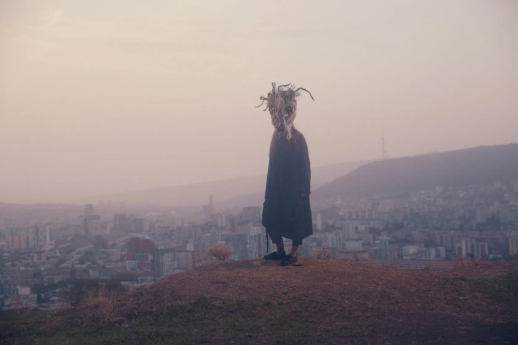 A figure in a shaggy mask and long dark robe stands on a hilltop above a hazy city.
