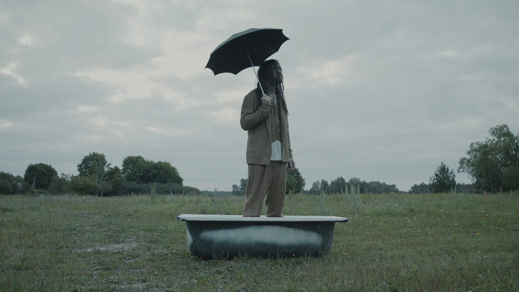 A man with an umbrella stands in an old bathtub in the middle of a field.