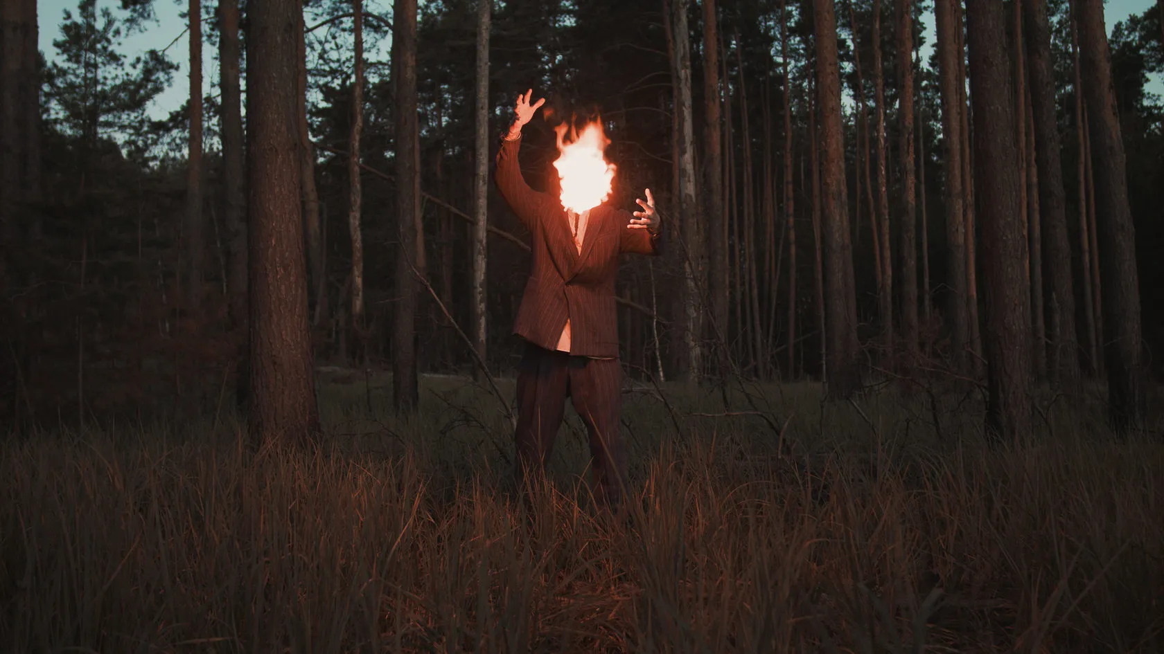 A man in a dark pine forest holds up his hands, his head a ball of fire.