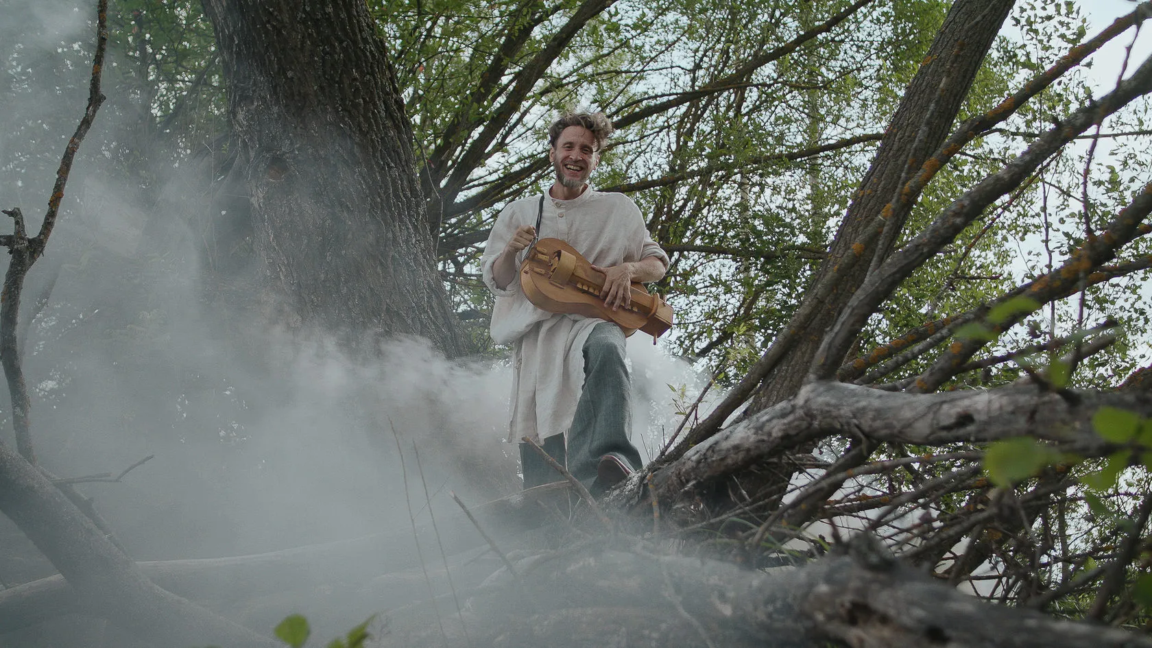 A laughing man in a linen shirt plays a hurdy-gurdy among fallen trees and smoke.