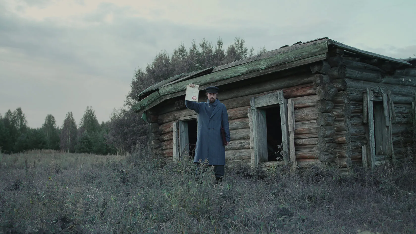 A man in a long coat and cap holds up a paper outside a crumbling log house.