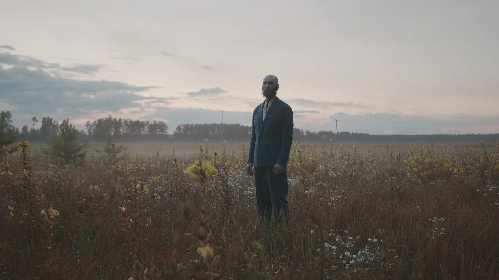 A bald man in a dark suit stands alone in a misty field under a pale sky.