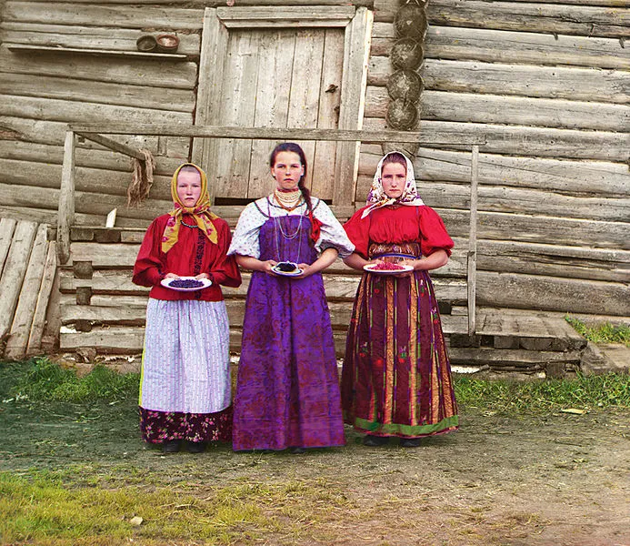 Three young women in folk dress offer berries outside a log house, a colour photograph by Prokudin-Gorskii.