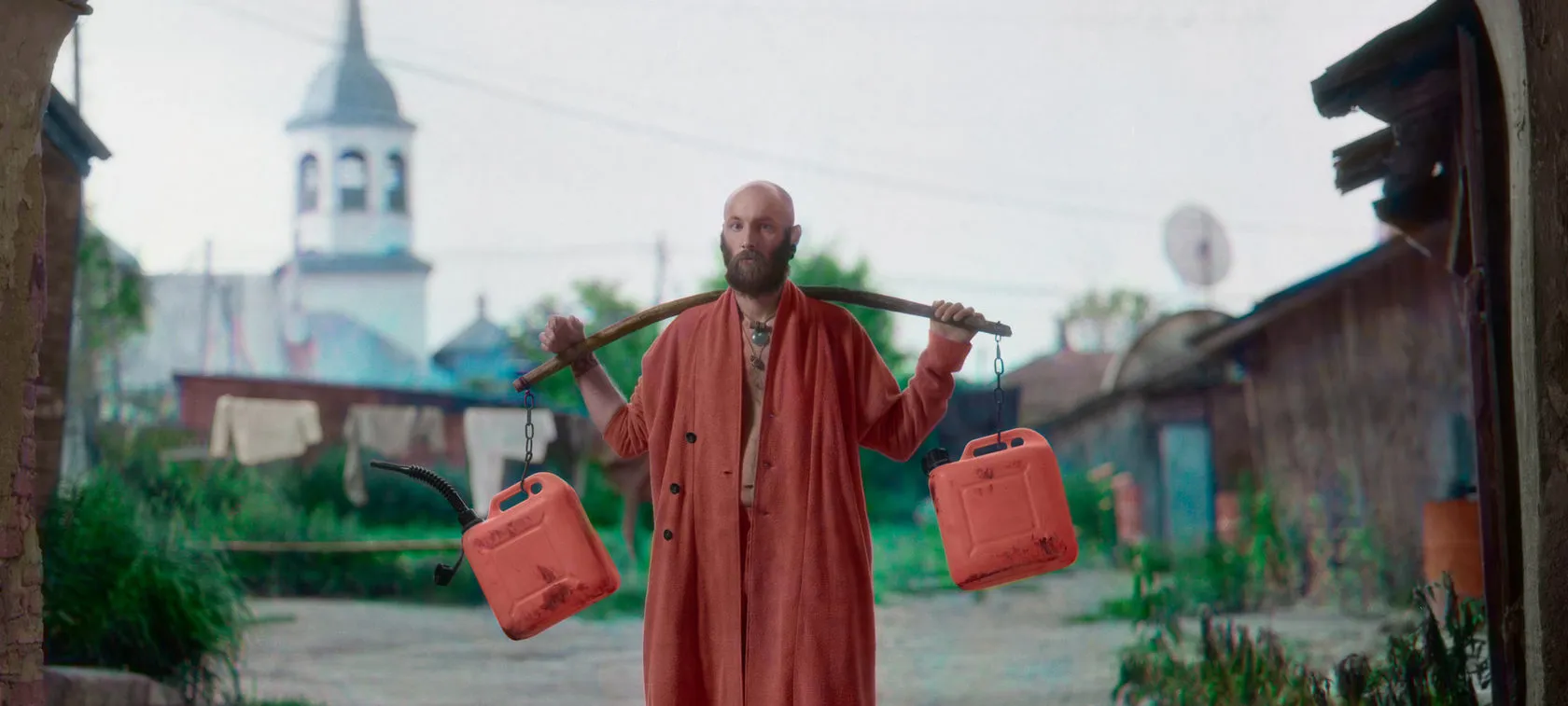 A bald, bearded man in a red robe carries two red fuel cans on a yoke, a bell tower behind.