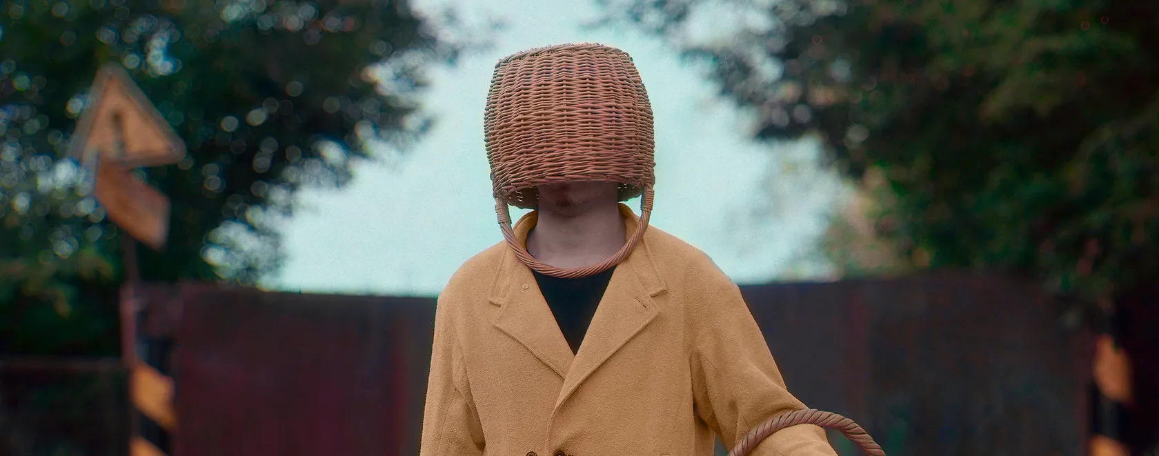 A man in a mustard suit with a wicker basket over his head stands by a red gate.