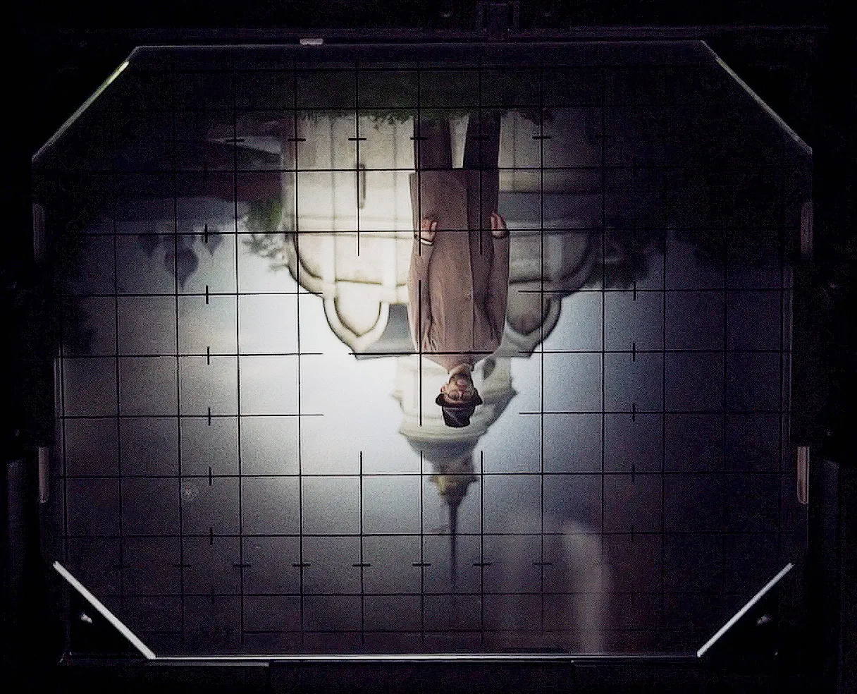 A model by a white church appears upside down on the gridded ground glass of a large-format camera.