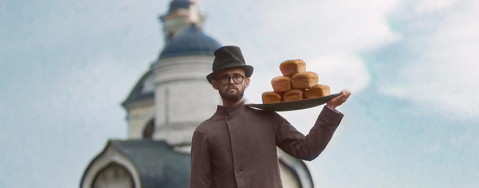 A man in a hat and glasses balances a tray of bread loaves on his shoulder by a church dome.