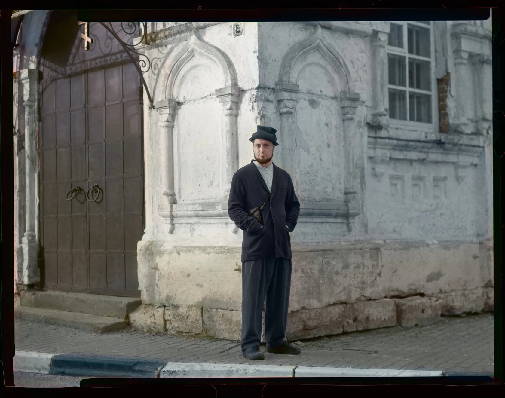 A man in a dark coat and hat stands by the carved white wall of an old building.