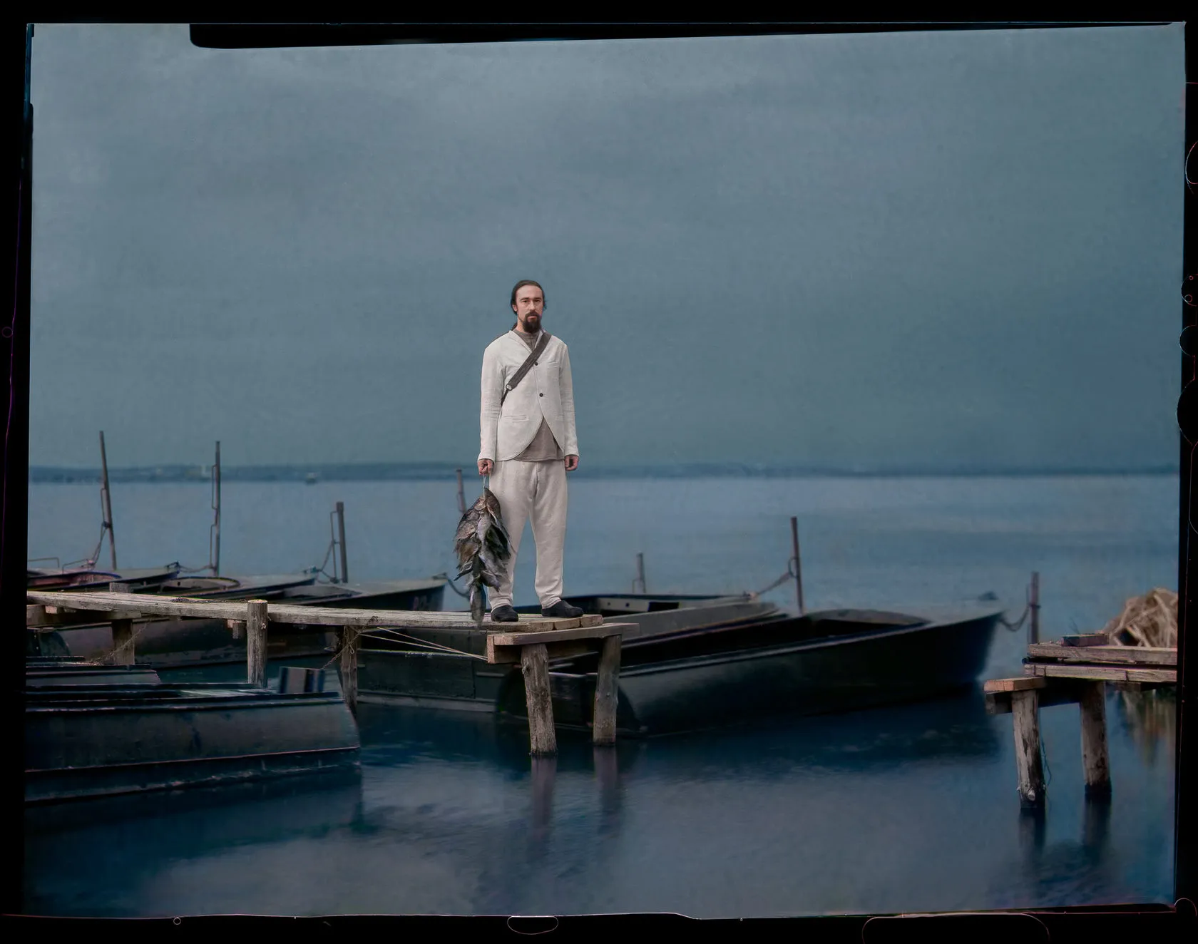 A man in a white suit stands on a wooden pier among moored boats on a grey lake.