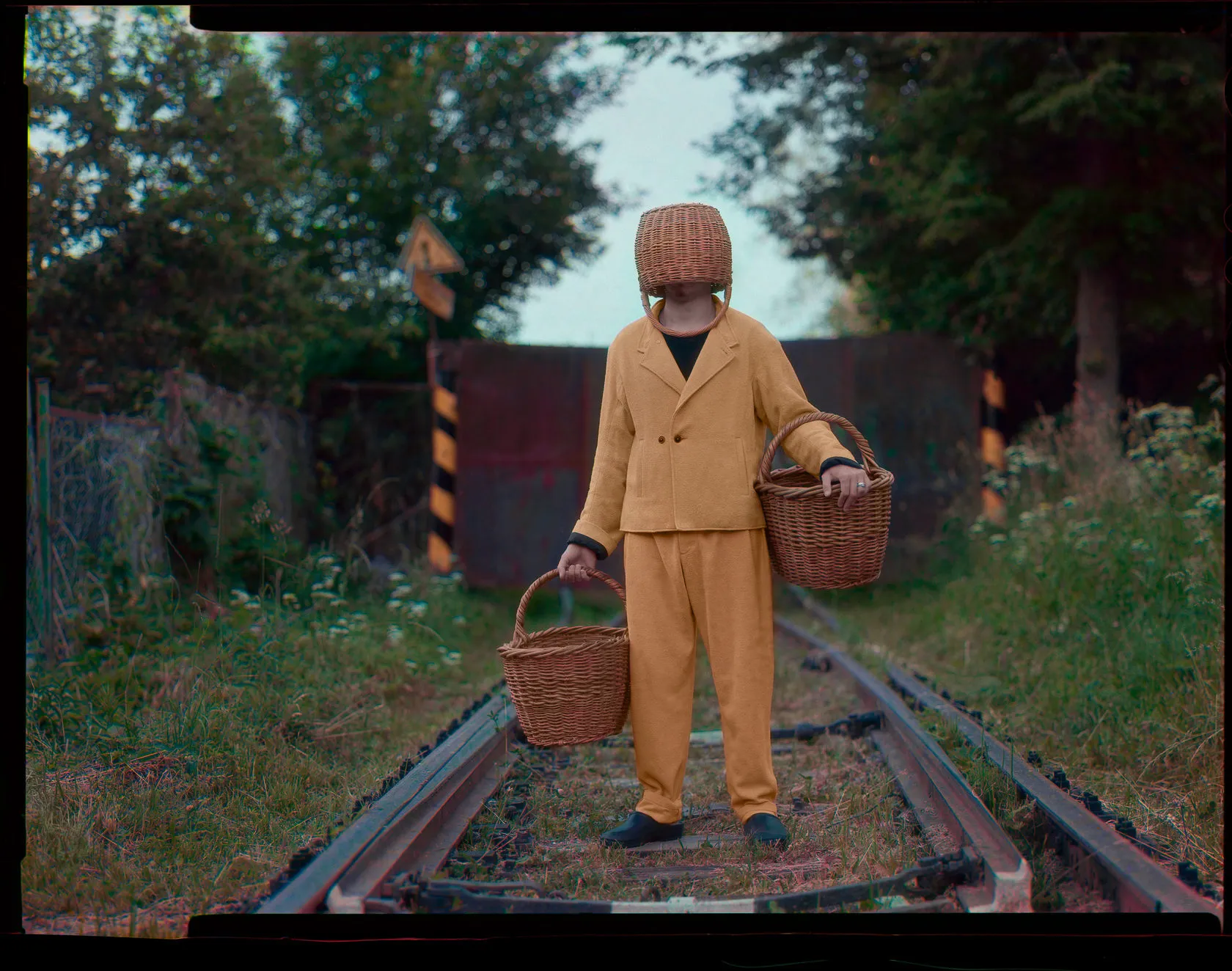 The basket-headed man in mustard stands on rail tracks, a wicker basket in each hand.