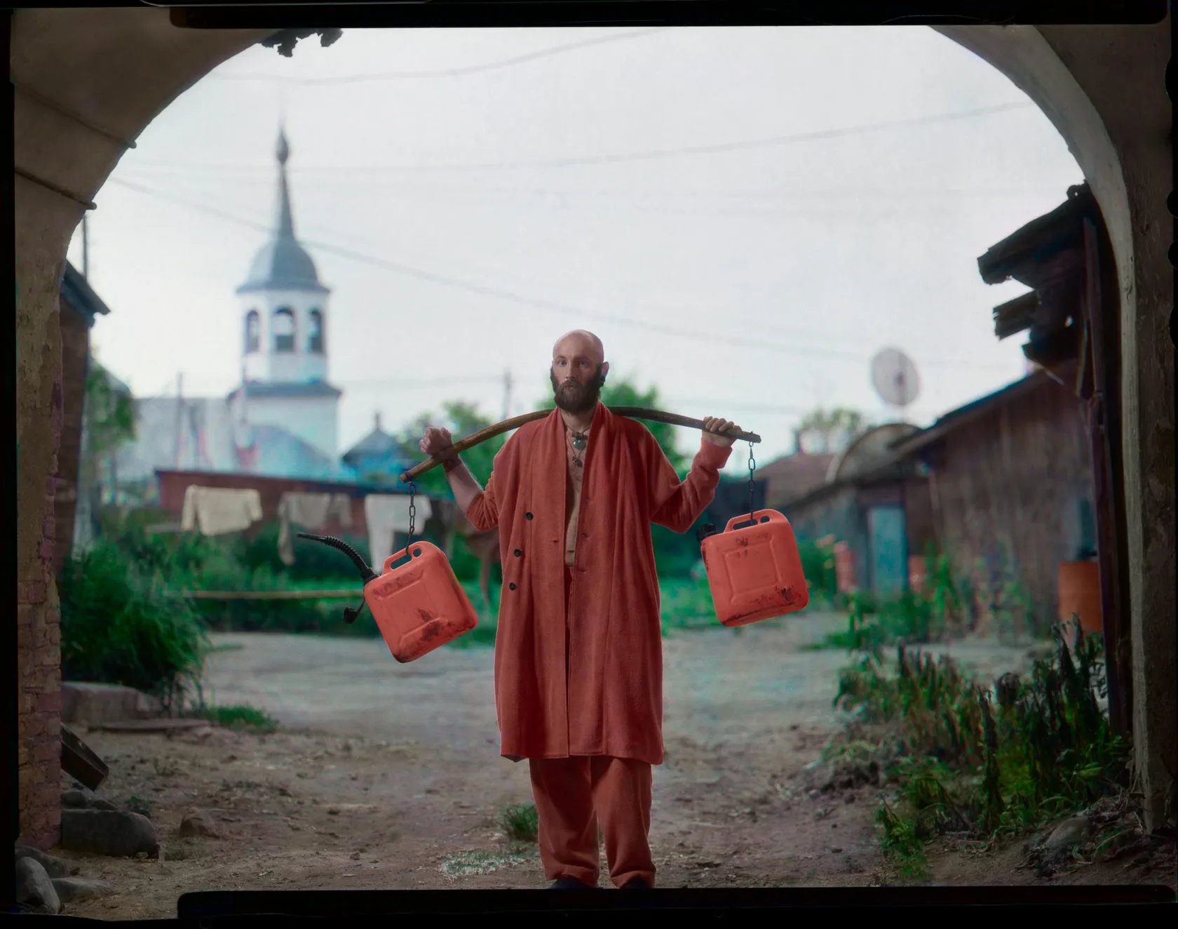 The man in the red robe carries fuel cans on a yoke under an archway, a bell tower behind.