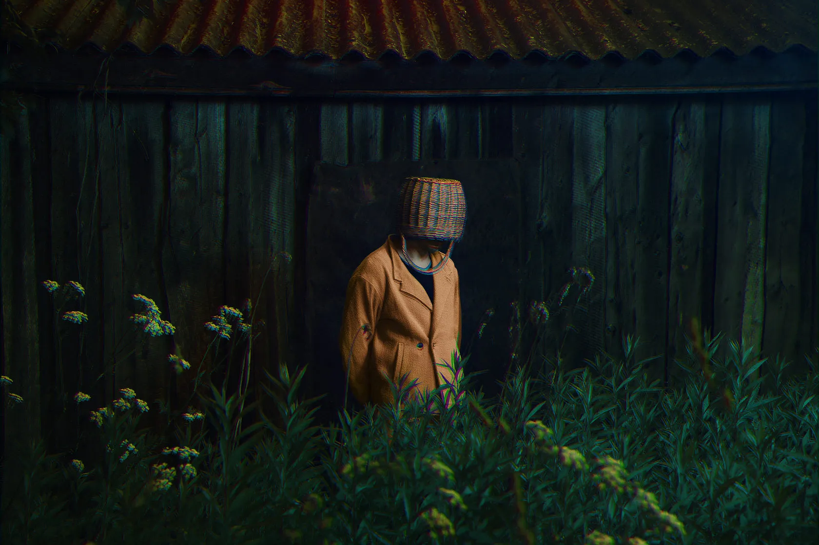 The basket-headed man in mustard stands in tall grass by a dark wooden shed.