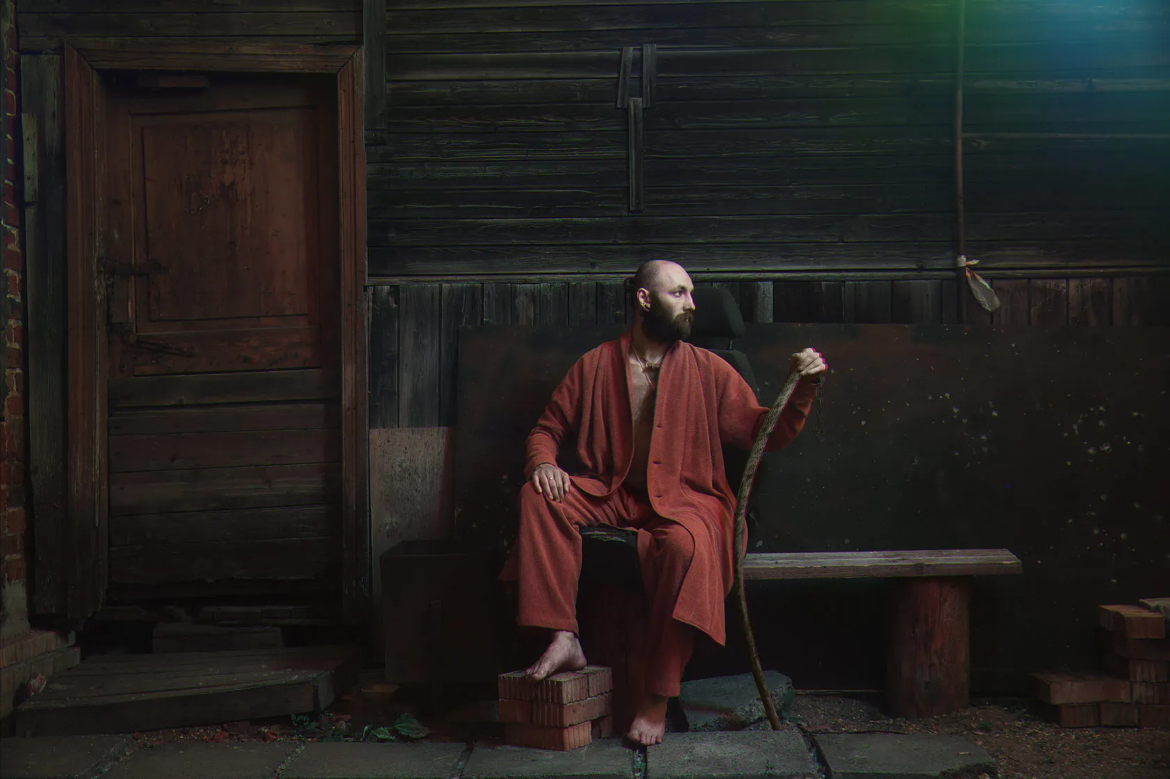 A barefoot man in a red robe sits on a bench by a wooden door, holding a staff.