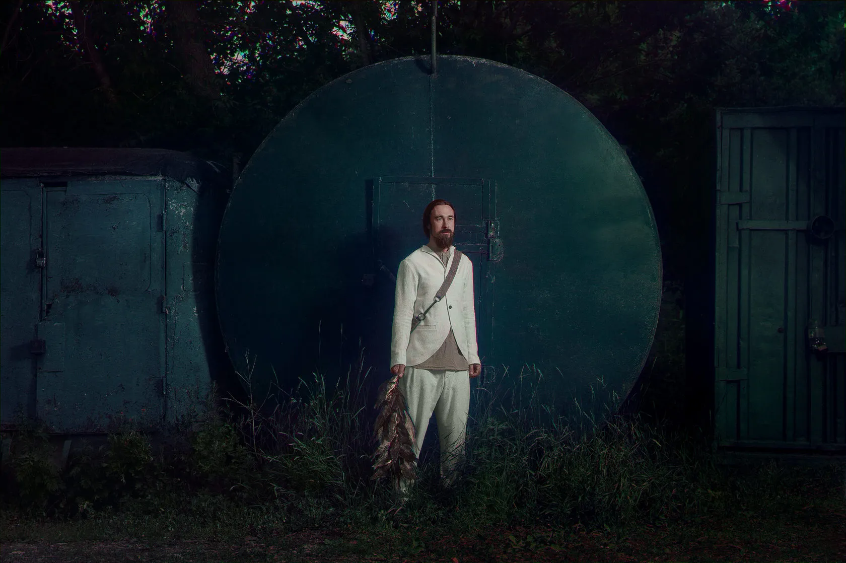 A man in a white suit holds a string of dried fish before a huge round green door.