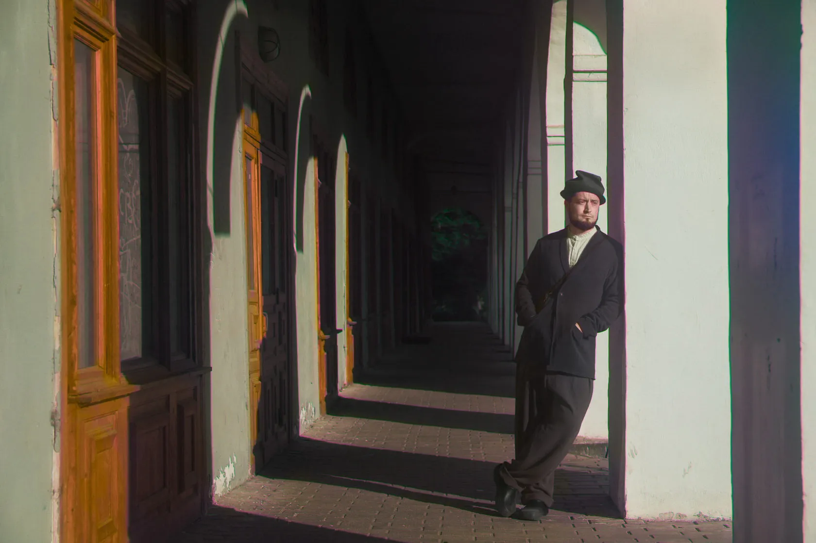 A man in a black suit and hat leans against a pillar in a shaded arcade.