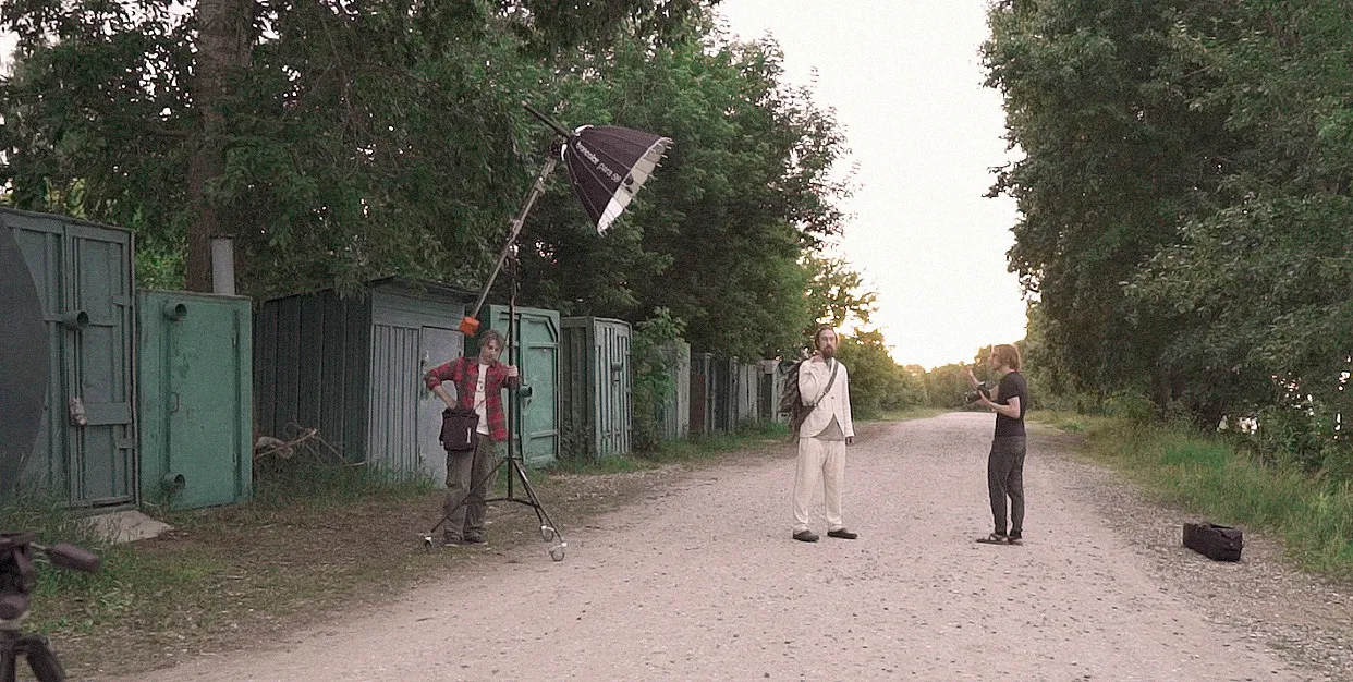 The crew sets a Broncolor light on a stand as a model in white waits on a gravel road.