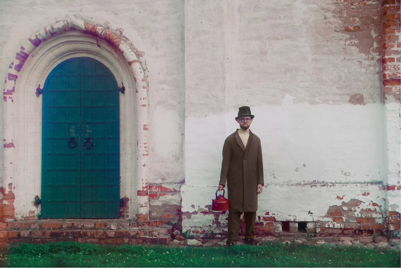 A man in a long brown coat and a hat stands by a whitewashed church wall and a green door, holding a red kettle.
