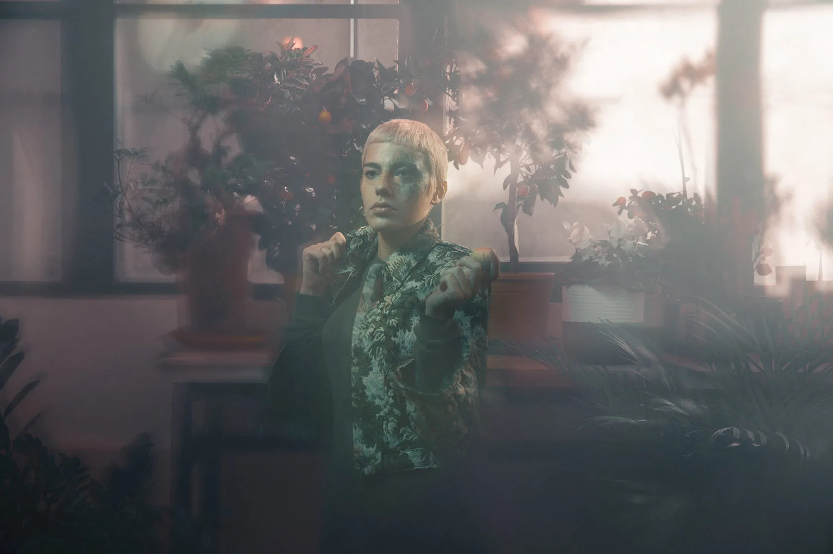 The model stands by a misty window among plants, hands at the collar of the floral jacket.