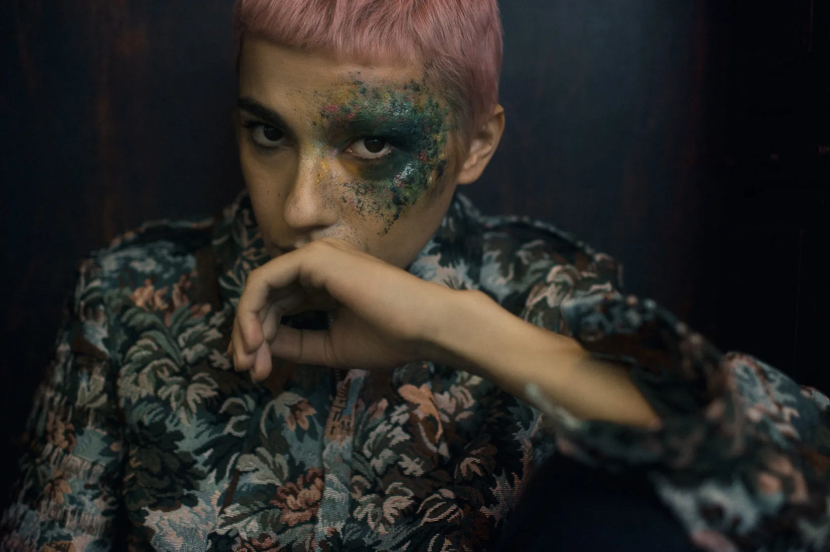 Close-up: the model rests her chin on her hand, green and gold glitter around one eye.