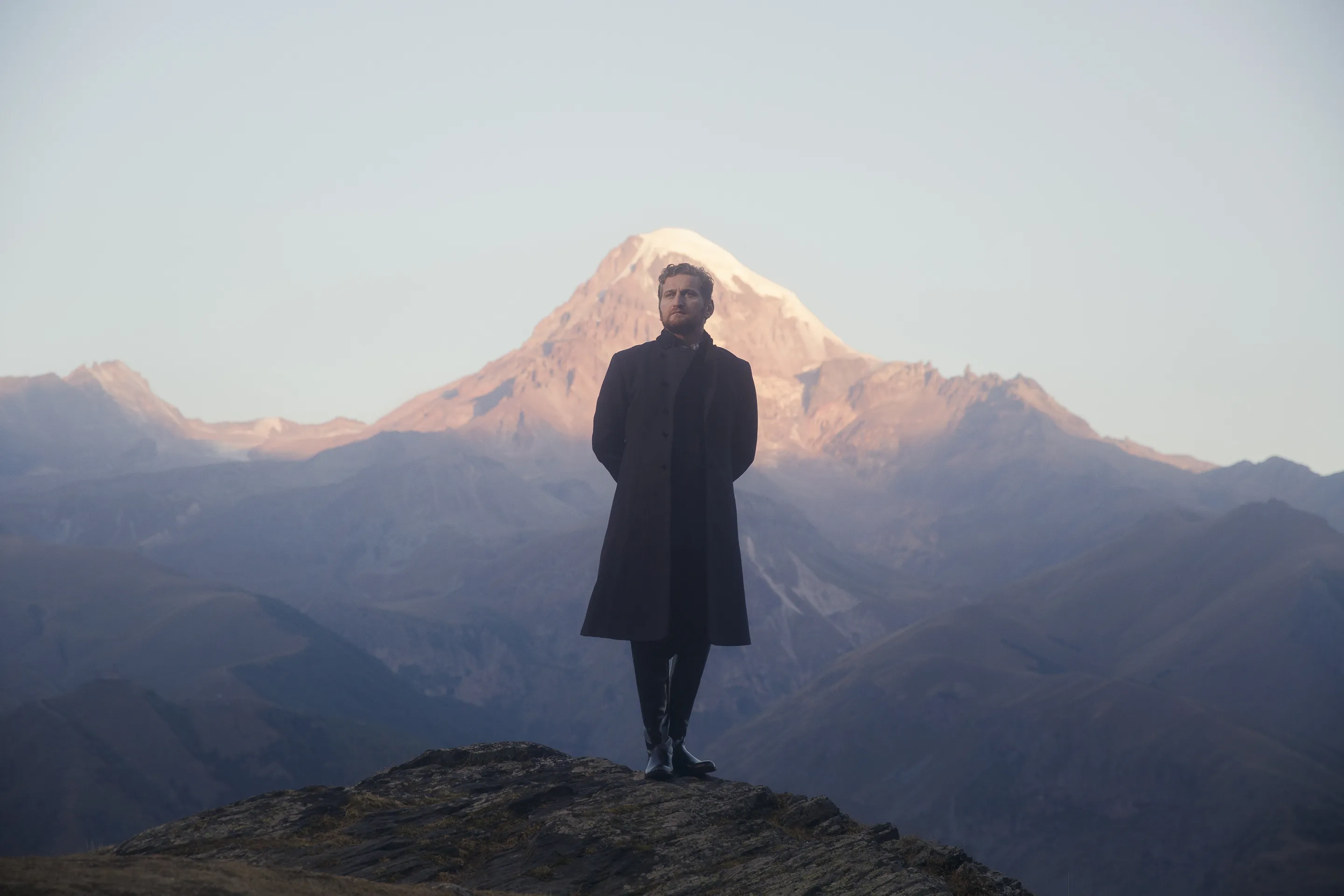 Jimsher campaign by Andrei Kovalev: a man in a dark coat stands on a rocky ridge facing a snow-capped peak at dawn.