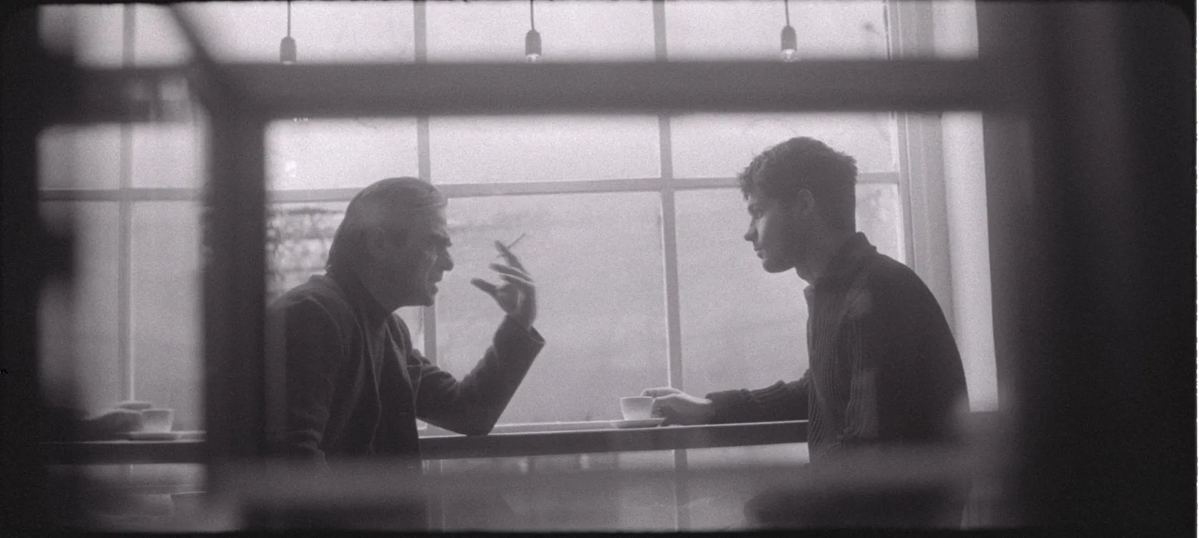 Seen through a café window, an older man with a cigarette talks to a young man over coffee.
