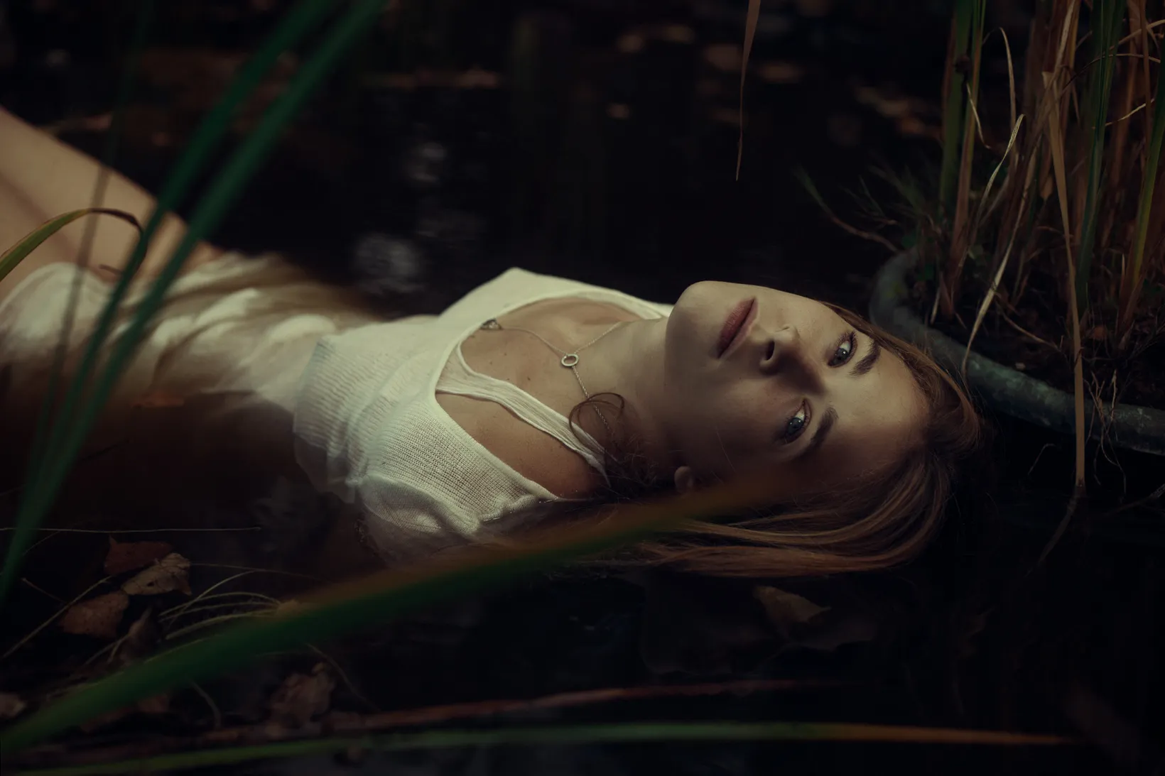 Emma Becker lies in dark water among reeds, eyes open, in a white dress.