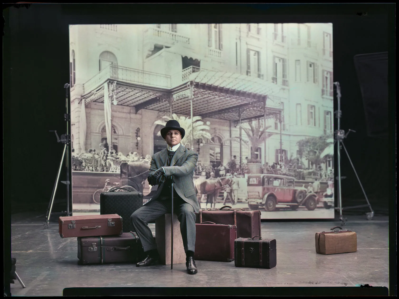 A man in a bowler hat sits on a pile of suitcases in front of a colour hotel backdrop.