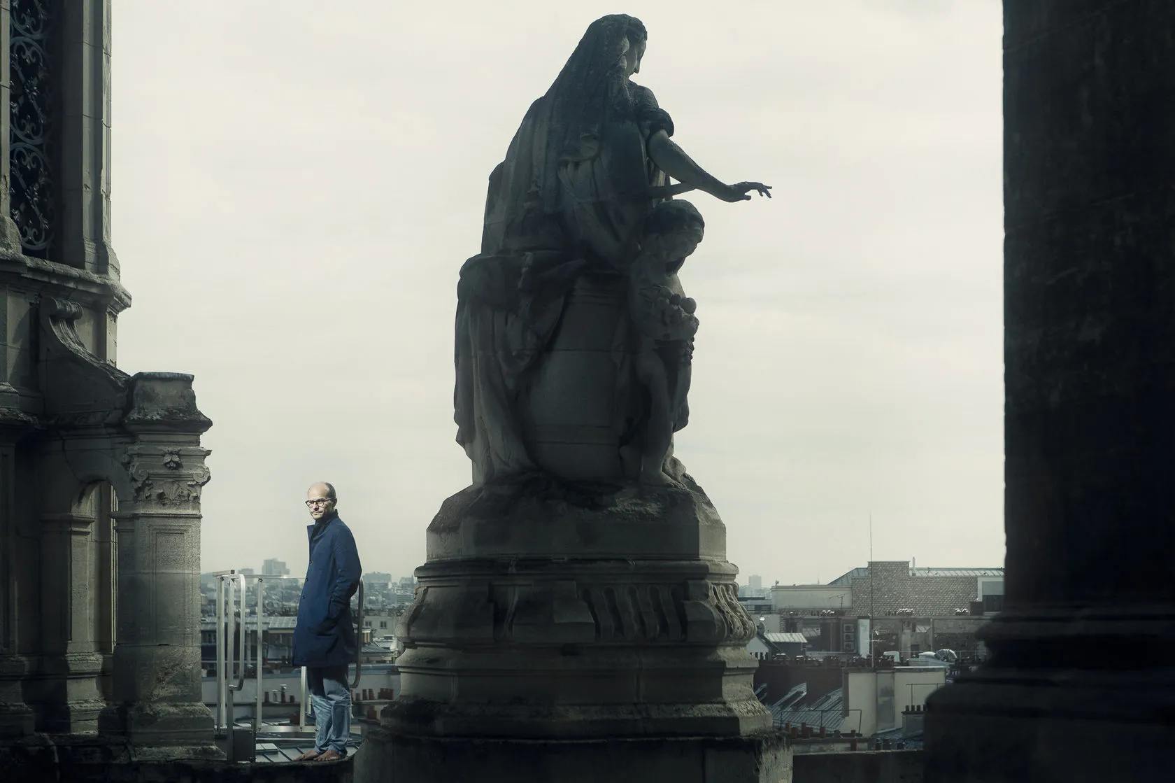 Laurent Clerc stands on a church roof beside a stone statue, Paris rooftops behind him.