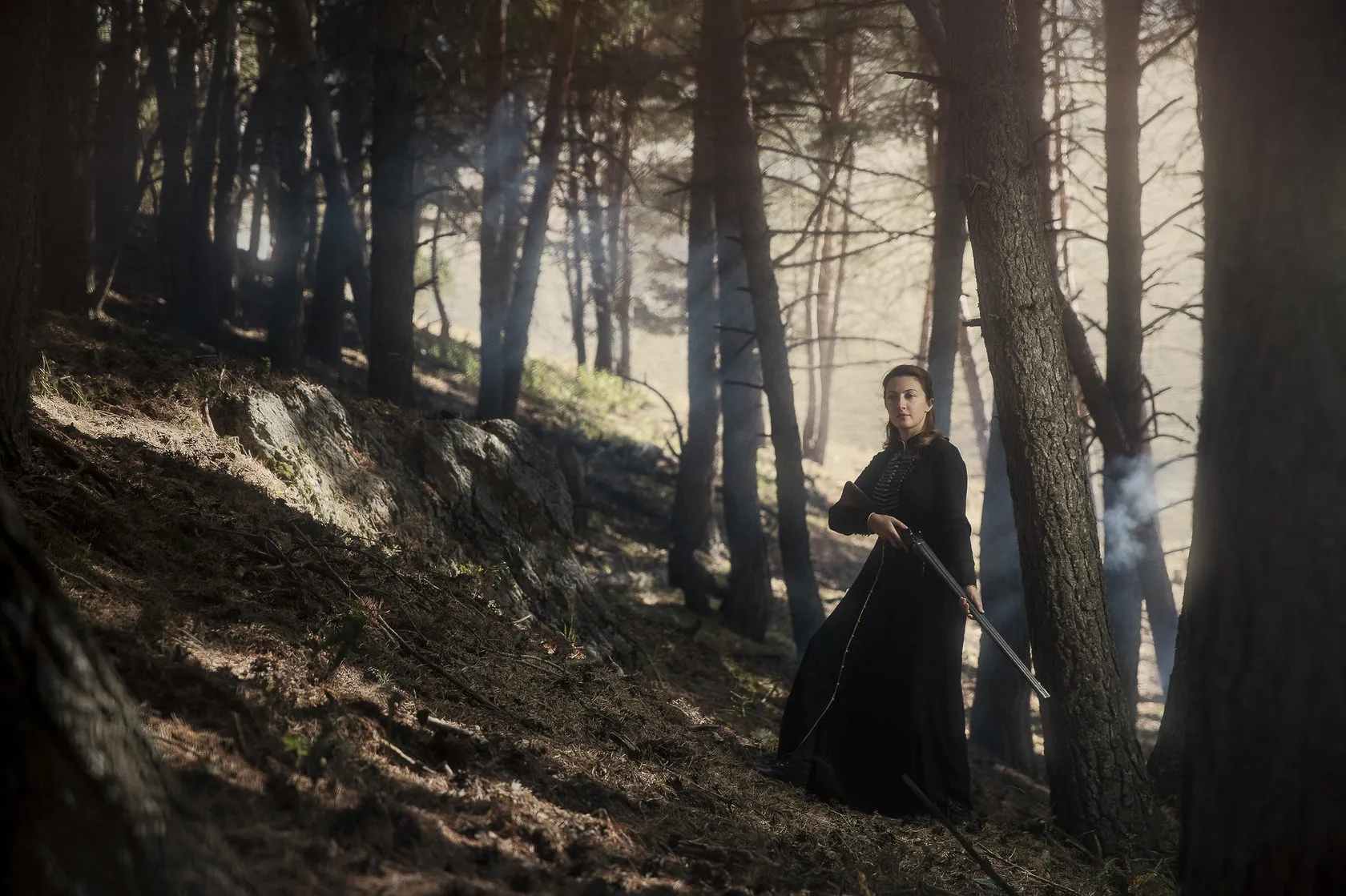 Marika stands on a pine forest slope in a long black dress, holding a shotgun.