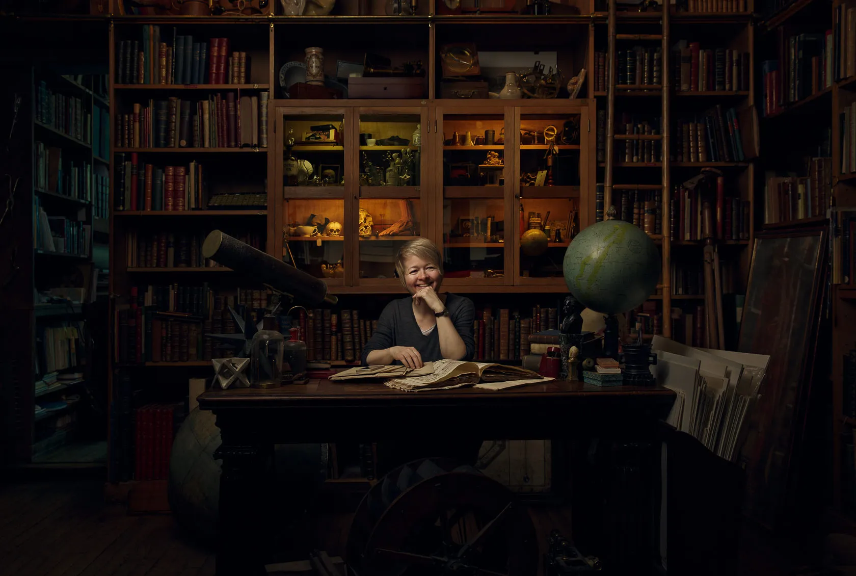 Sarah Waters sits at a desk in a book-lined study, a globe beside her.