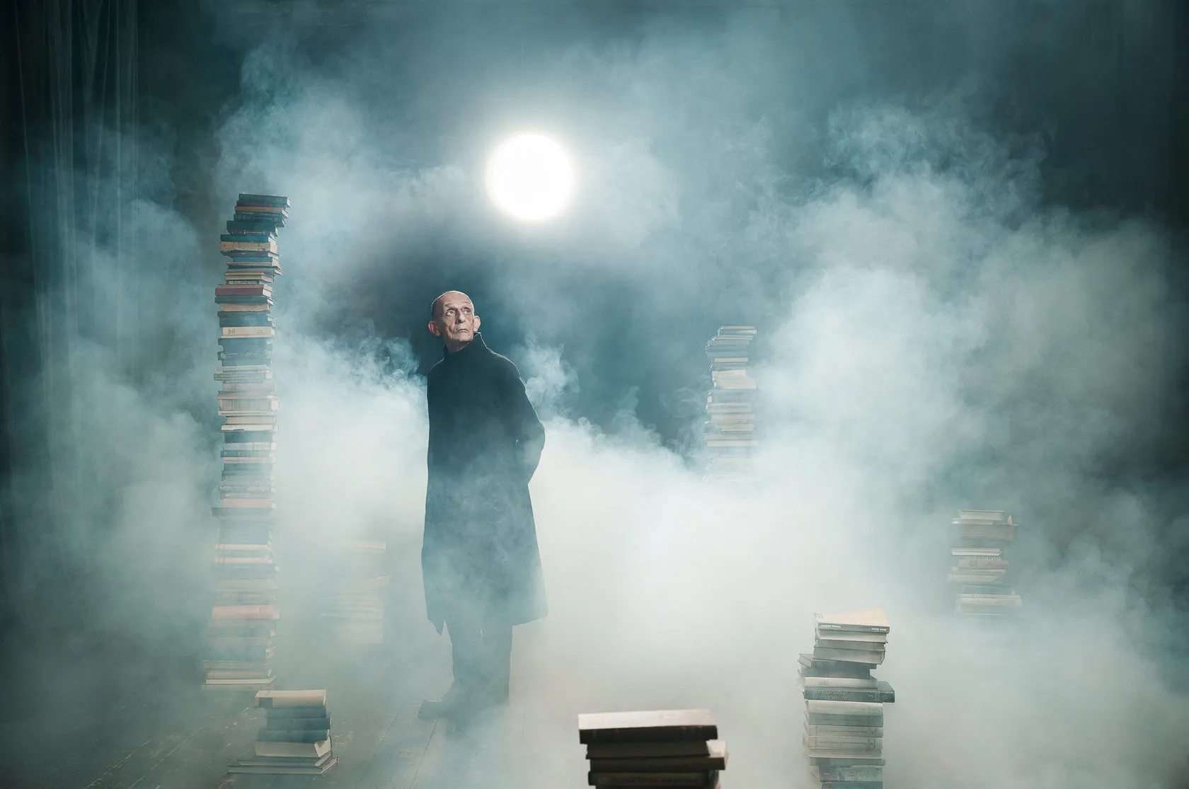 Victor Golyshev stands in smoke between towers of stacked books, a pale light above.
