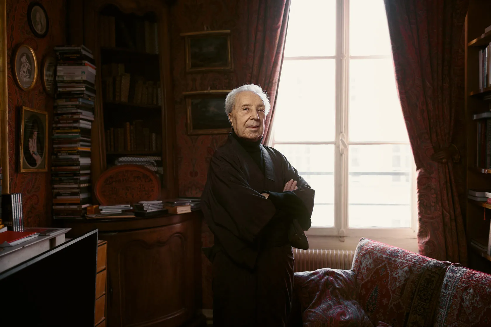 Marc Fumaroli stands with his arms folded by a window in a study full of books.