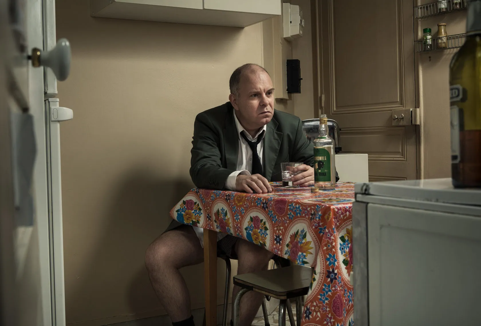Olivier Maulin sits bare-legged at a kitchen table in a jacket and loose tie, a bottle beside him.