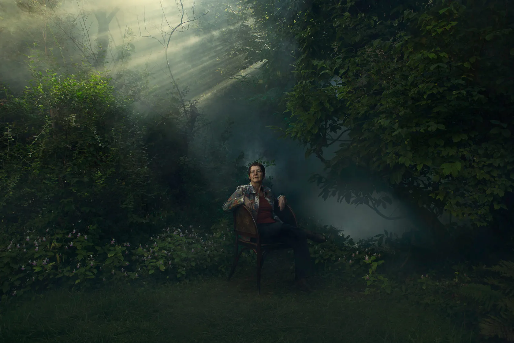 Jocelyne Saucier sits in a wicker chair in a misty garden, sunbeams falling through the trees.