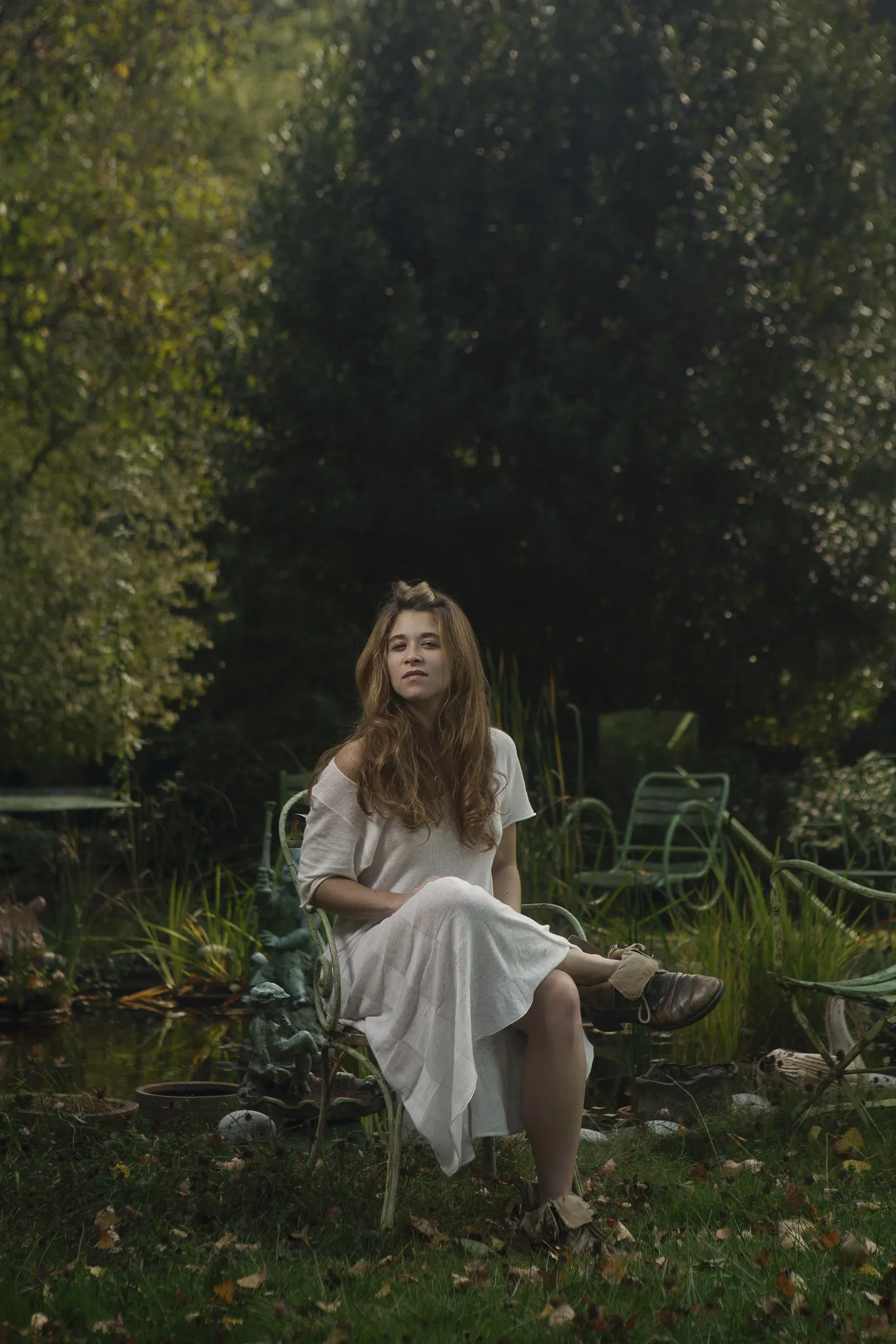 Emma Becker sits on a garden chair in a white dress, dark trees behind her.