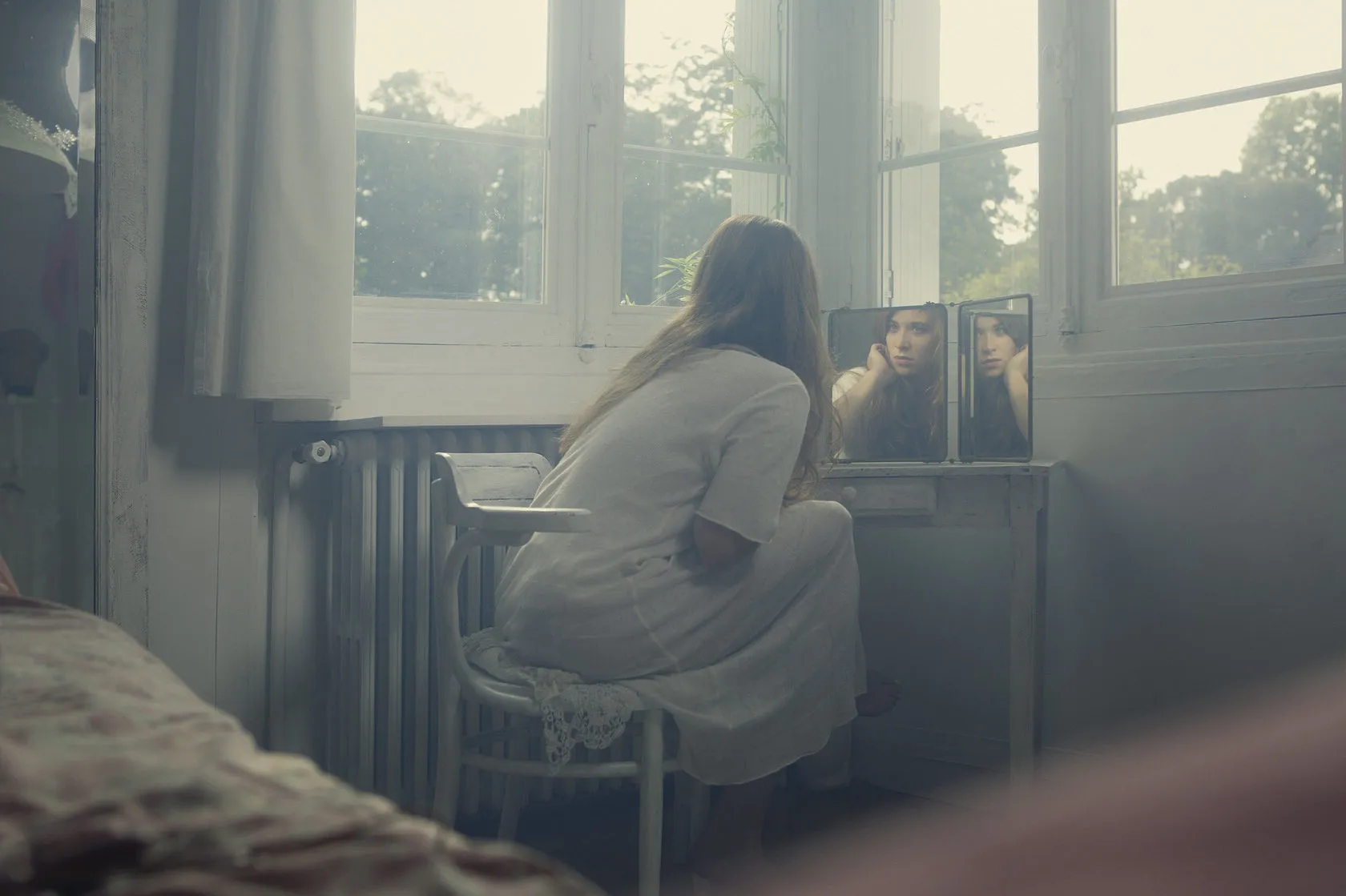Emma sits at a dressing table by tall windows, her face caught in a folding mirror.