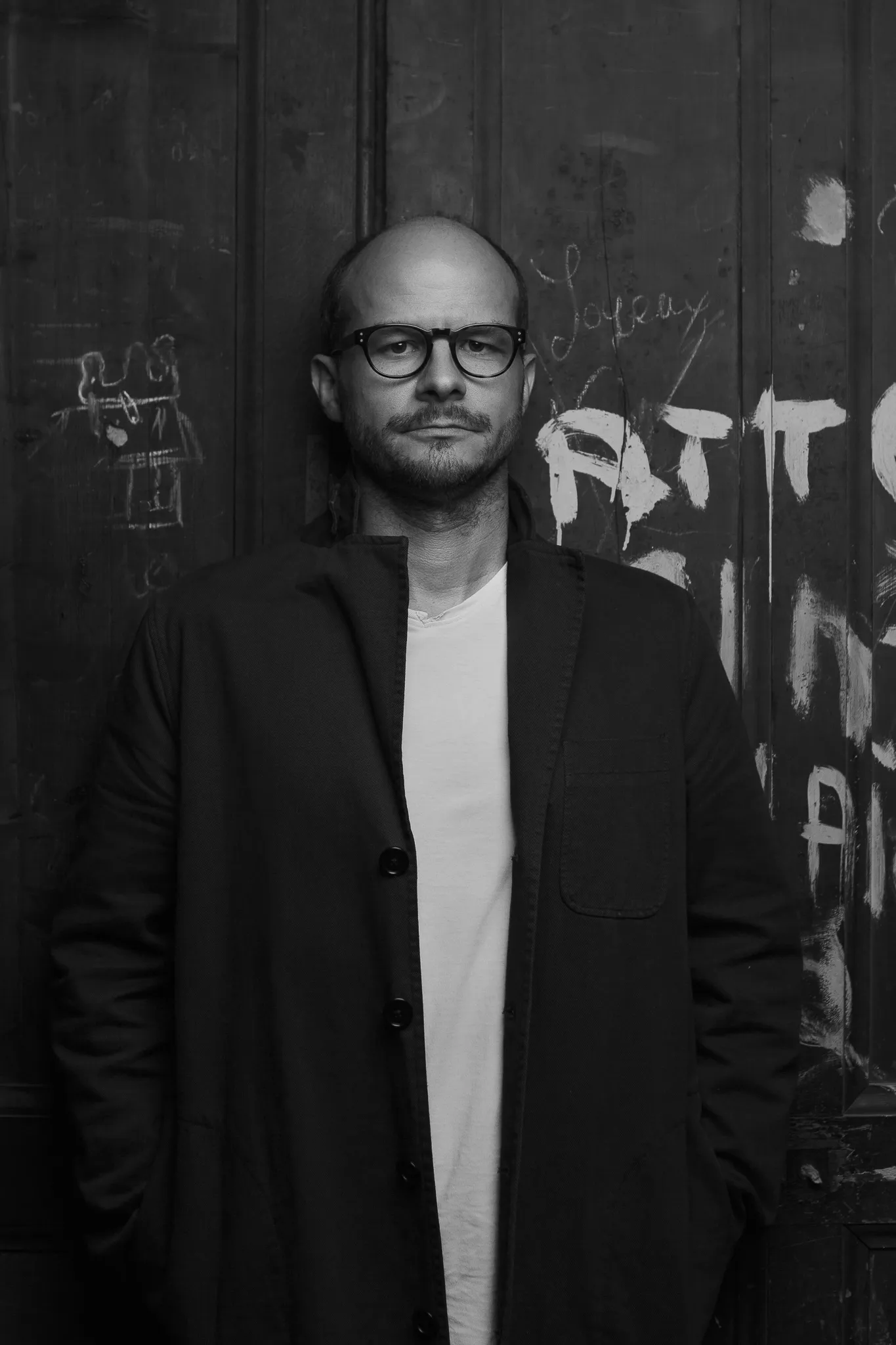 Laurent in glasses and a dark coat against a wall covered in graffiti, in black and white.