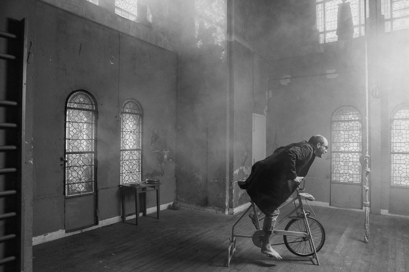 Laurent pedals an exercise bike in a smoky, empty hall with arched windows.