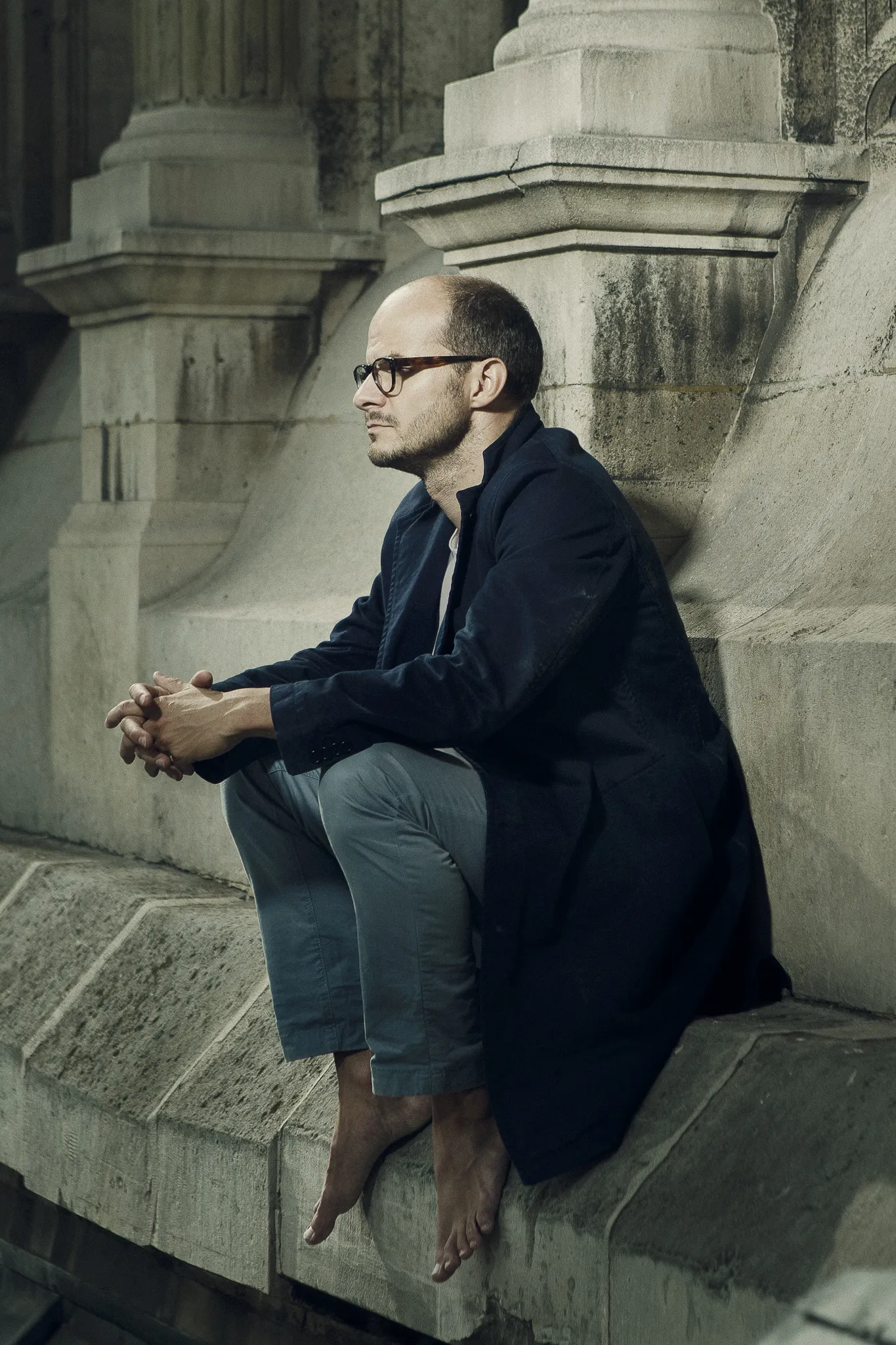 Laurent sits barefoot on a stone ledge, his hands clasped.