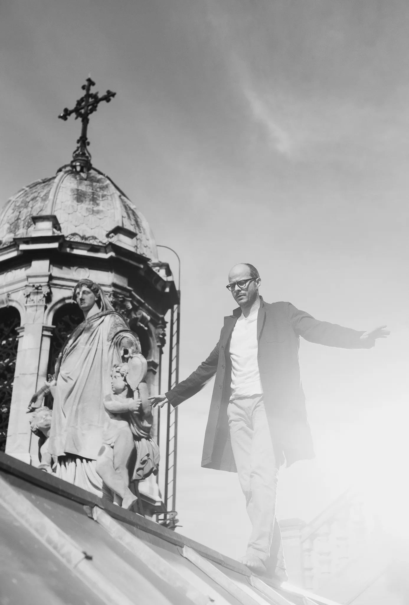 Laurent balances on the roof with his arms out, beside a dome topped by a cross.