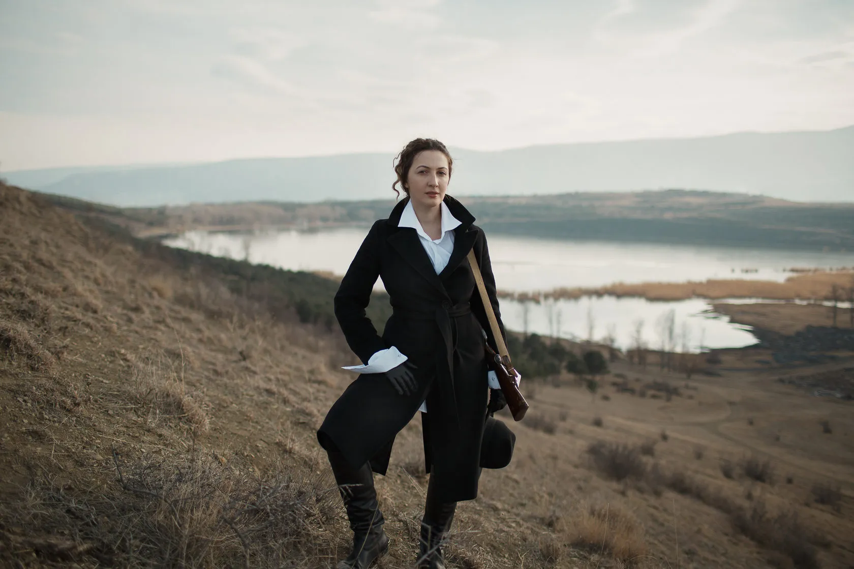 Marika in a long black coat and white shirt stands on a hillside above a lake.