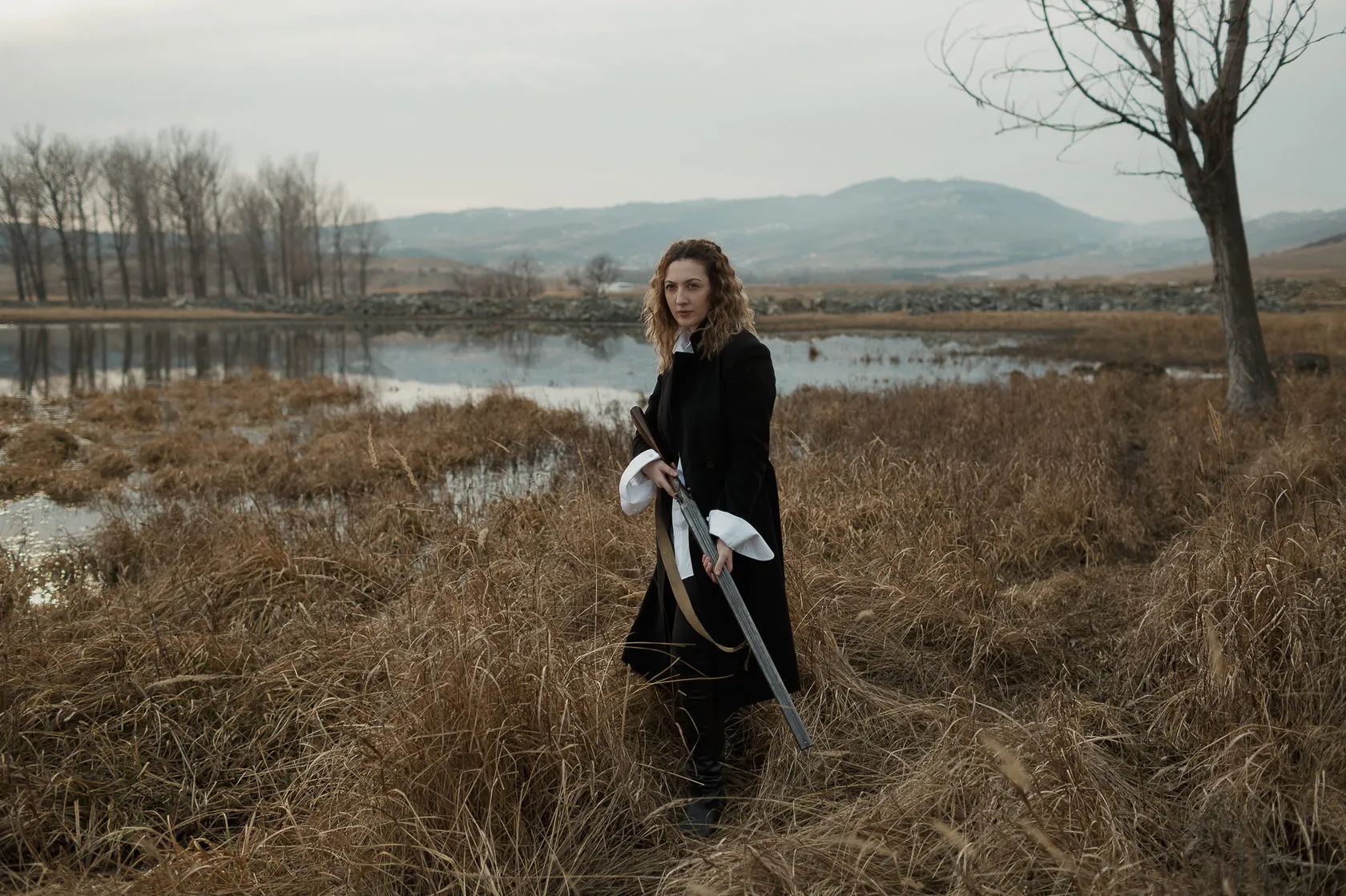 Marika walks through tall dry grass by a lake, a shotgun in her hand.