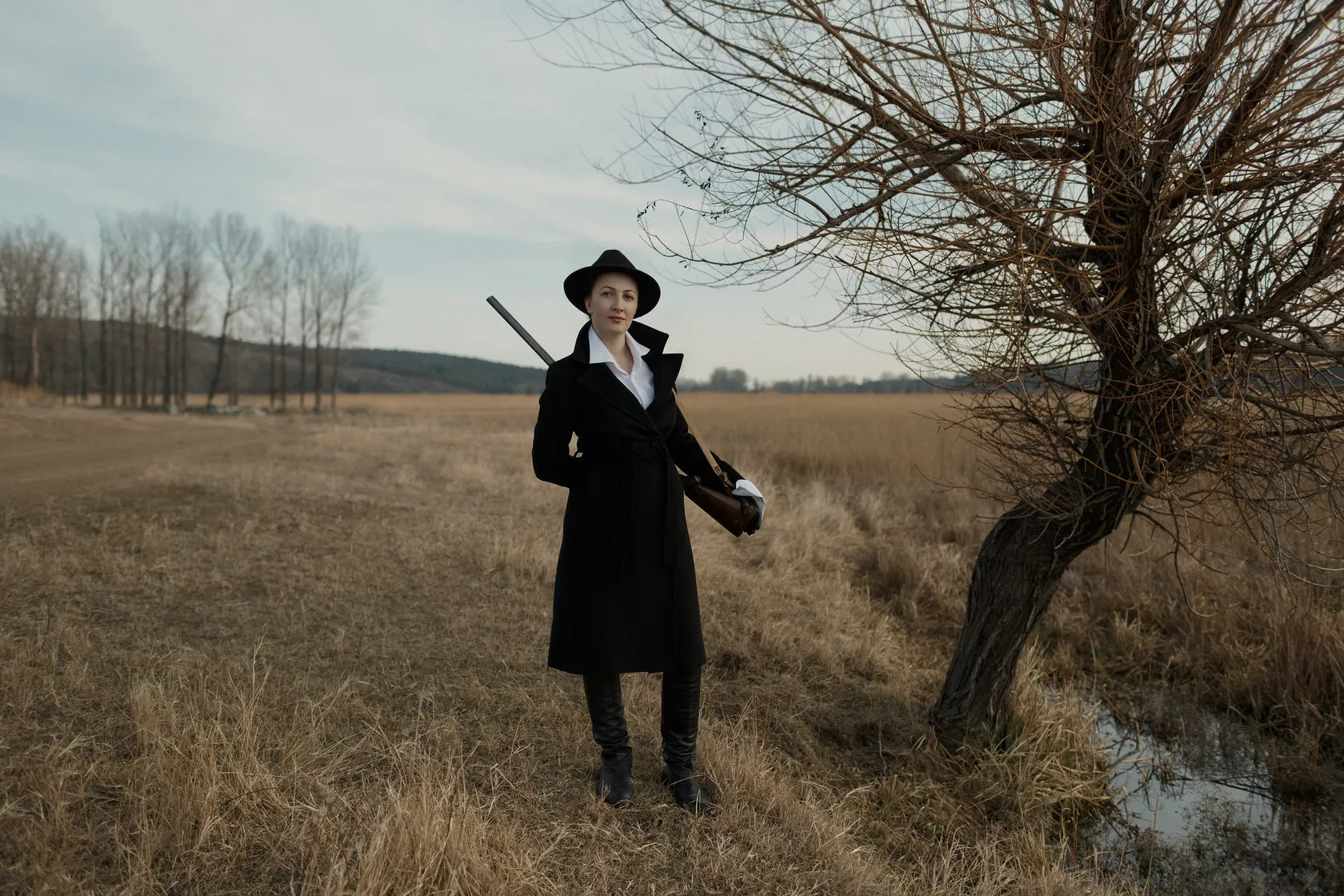 Marika in a black hat and coat stands in a field by a bare tree, a gun on her shoulder.