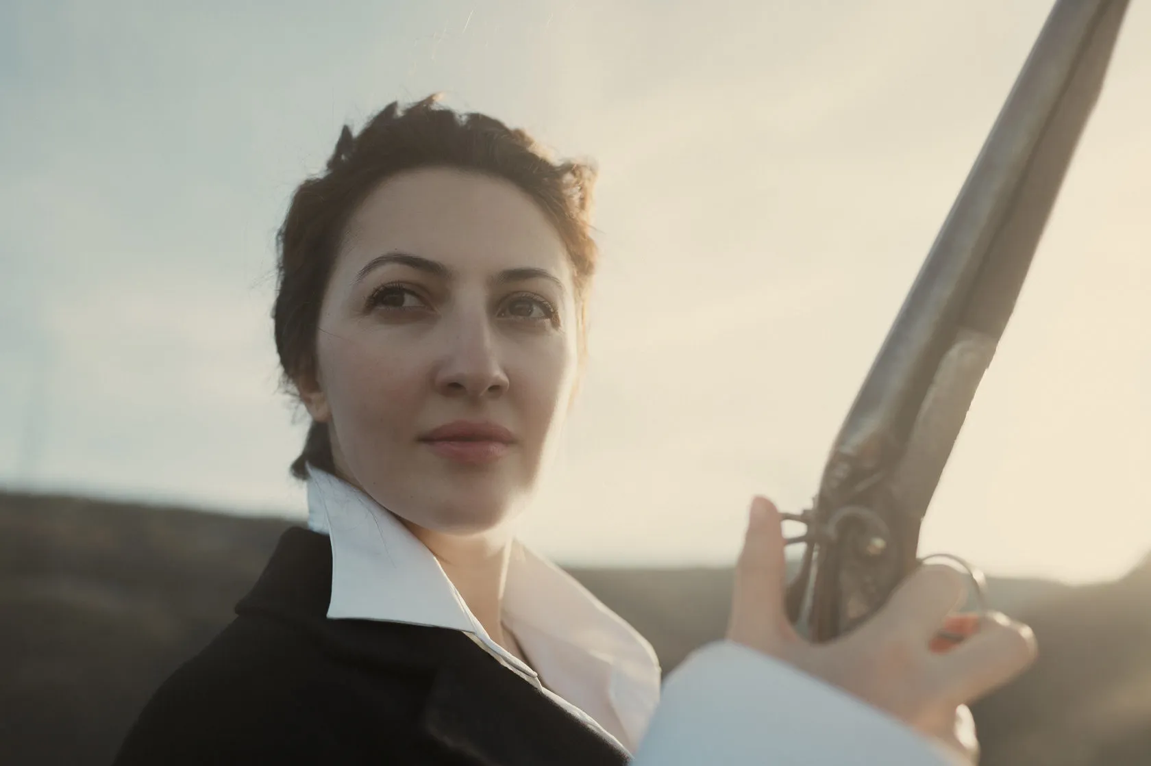 Marika holds a shotgun upright, close up, in soft evening light.